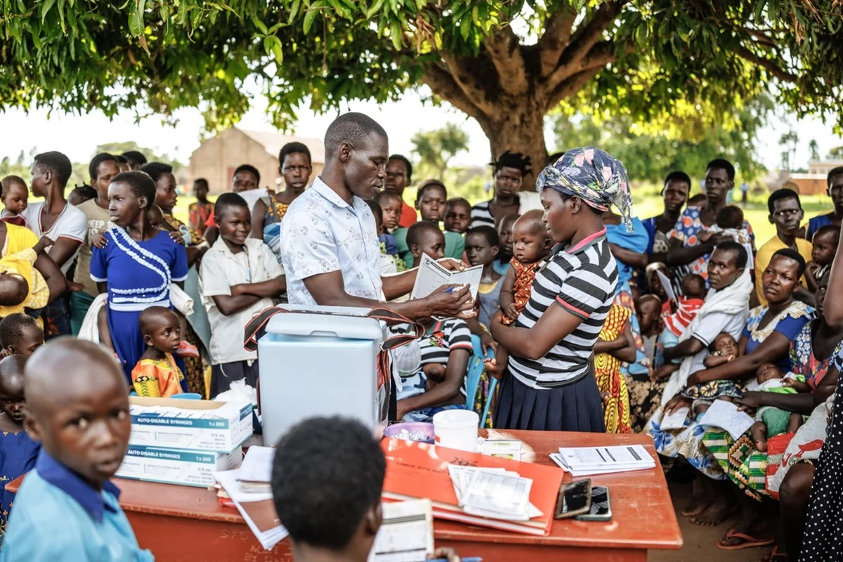 2025, Distrikt Apac, Uganda.  Im Rahmen eines Community-Outreachs erklärt ein Gesundheitshelfer einer Mutter, dass der R21/Matrix-M-Malariaimpfstoff in vier Dosen verabreicht wird – im Alter von 6, 7, 8 und 18 Monaten. Mit Unterstützung von WHO, Gavi, UNICEF und weiteren Partnern ist Uganda das 19. afrikanische Land, das den Malariaimpfstoff in sein reguläres Impfprogramm aufgenommen hat. Der offizielle Start im Distrikt Apac im Norden Ugandas markierte den bislang größten Rollout und richtet sich an über 105 Bezirke mit hoher und mittlerer Übertragungsrate.